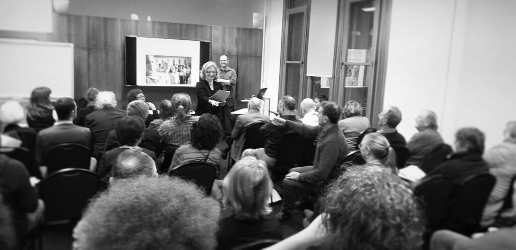 Black and white image of LHP AGM, The audience is shown from behind, looking at Cybèle Locke who is presenting.