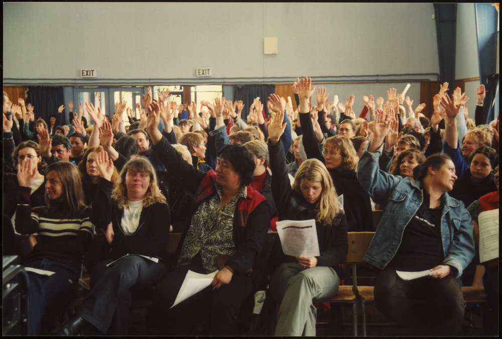 A colour image of  a group, mostly women, voting. We can see papers in the hands of people in the front row.